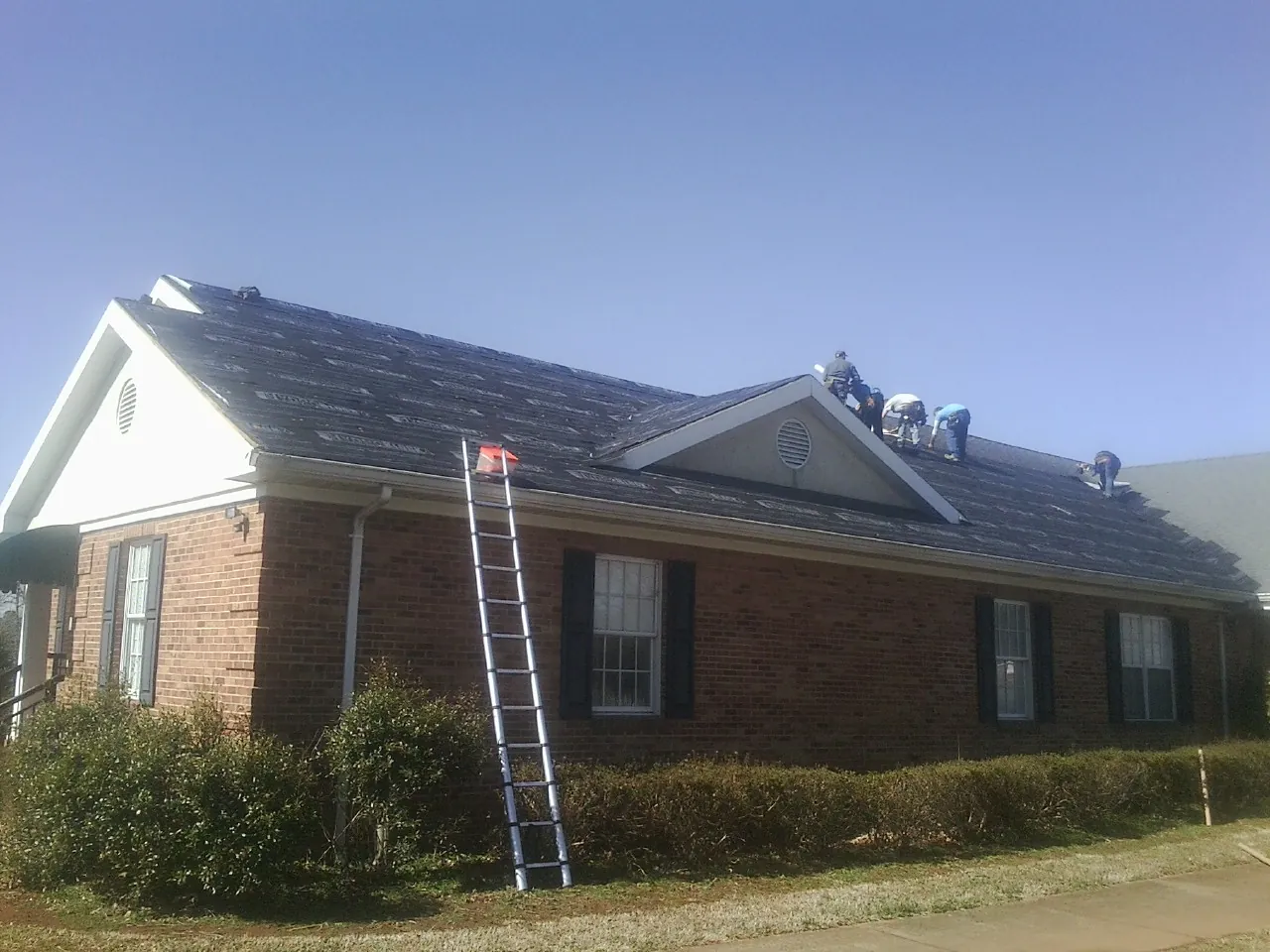 Crew tearing shingles off a brick ranch roof with a ladder set — Metal Roof Repair work by our crew in Bayport