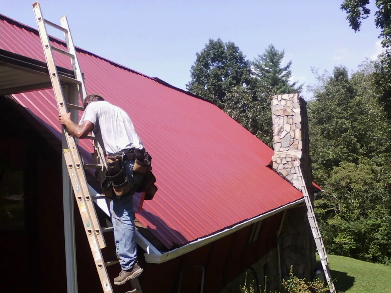 Roofer on a ladder beside a red metal roof and stone chimney — Soffit & Fascia Repair by Roofing Brothers in Bayport